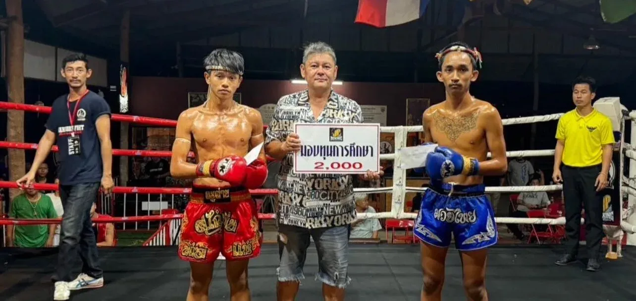 Two young Muay Thai fighters stand in a boxing ring with their trainer holding a 2000 Baht check